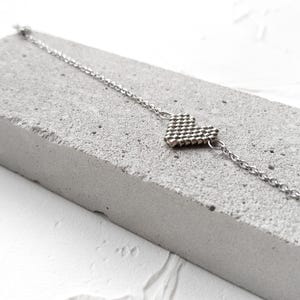 Op de afbeelding: Een zilverkleurige armband met een centraal hartvormig bedeltje, opgebouwd uit kleine, uniforme metalen kraaltjes. Het bedeltje is bevestigd aan een fijne, dunne zilverkleurige ketting met kleine ronde schakels aan beide zijden.