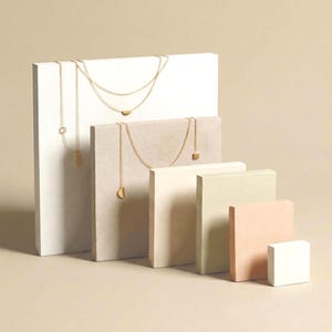May include: Two gold necklaces with delicate chains and geometric pendants displayed on rectangular blocks of varying heights and neutral colors. One necklace features a semi-circular charm, while the other includes a small square and a semi-circle.