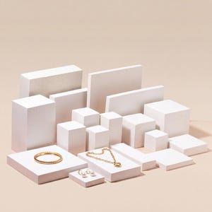 Puede incluir: Un brazalete de metal trenzado en tono dorado, un collar de cadena dorada con un colgante en forma de corazón y un par de pendientes pequeños dorados con detalles esféricos blancos, sobre bloques rectangulares blancos.