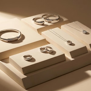 Puede incluir: Una colección de joyas de plata expuestas sobre plataformas beige. El surtido incluye anillos, pulseras y collares con colgantes y diseños circulares. Las joyas están dispuestas para mostrar los diferentes estilos y diseños.