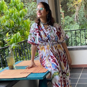 Puede incluir: Una mujer lleva un vestido maxi blanco, rojo, azul y amarillo con estampado ikat, cuello en V y mangas cortas. Está de pie en un porche de madera con una mesa y un taburete azules.