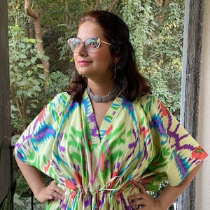 Op de afbeelding: Een vrouw draagt een lichtgroene kaftan met een kleurrijk ikat-patroon. De kaftan heeft een V-hals en een tailleband. De vrouw draagt een bril en een zilveren ketting.