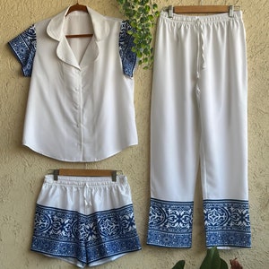 Op de afbeelding: Witte pyjamaset met een kort, geknoopt overhemd met korte mouwen, een lange broek en bijpassende shorts. Het overhemd en de broek zijn effen wit, terwijl de shorts een blauwe en witte geprinte rand hebben.