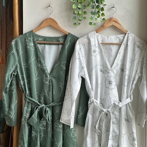 May include: Two floral print robes, one green and one white, hanging on wooden hangers. Both robes have a V-neckline and a tie belt.
