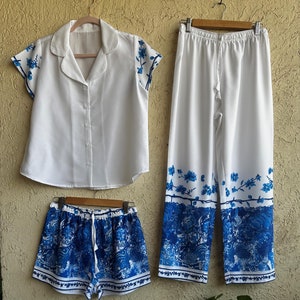 Op de afbeelding: Een witte pyjamaset met blauwe bloemenprint. De set bevat een top met korte mouwen en een knoopsluiting, een short en een broek. De broek heeft een trekkoord.