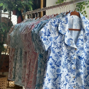 Può includere: Una fila di camicie bianche a maniche corte con motivi floreali blu appese a grucce. Le camicie hanno un colletto e una chiusura con bottoni.