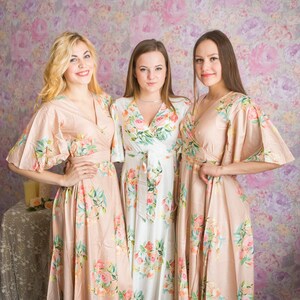 May include: Three women wearing long floral print robes. The robes are in shades of pink, white, and green. The women are standing in front of a pink and white floral patterned wall.