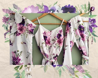 Puede incluir: Tres blusas blancas de estampado floral con flores moradas y verdes. Las blusas están colgadas en una percha de madera sobre un fondo verde.