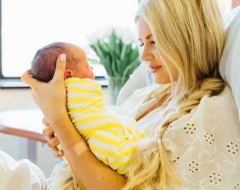 May include: A woman in a white dress with eyelet lace holds a newborn baby wrapped in a yellow and white striped blanket. The baby is looking up at the woman, who is smiling.