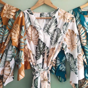 May include: Three floral print robes hanging on wooden hangers. The robes feature a monstera leaf pattern in shades of gold, white, and teal. Each robe has a wrap-around design with a tie closure at the waist.