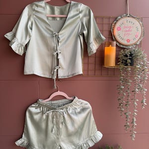 May include: A pale green satin pyjama set, comprising a top with a tie closure and frilled sleeves, and matching shorts with a drawstring waist and frilled hem. The set is displayed on pink hangers against a pink wall.