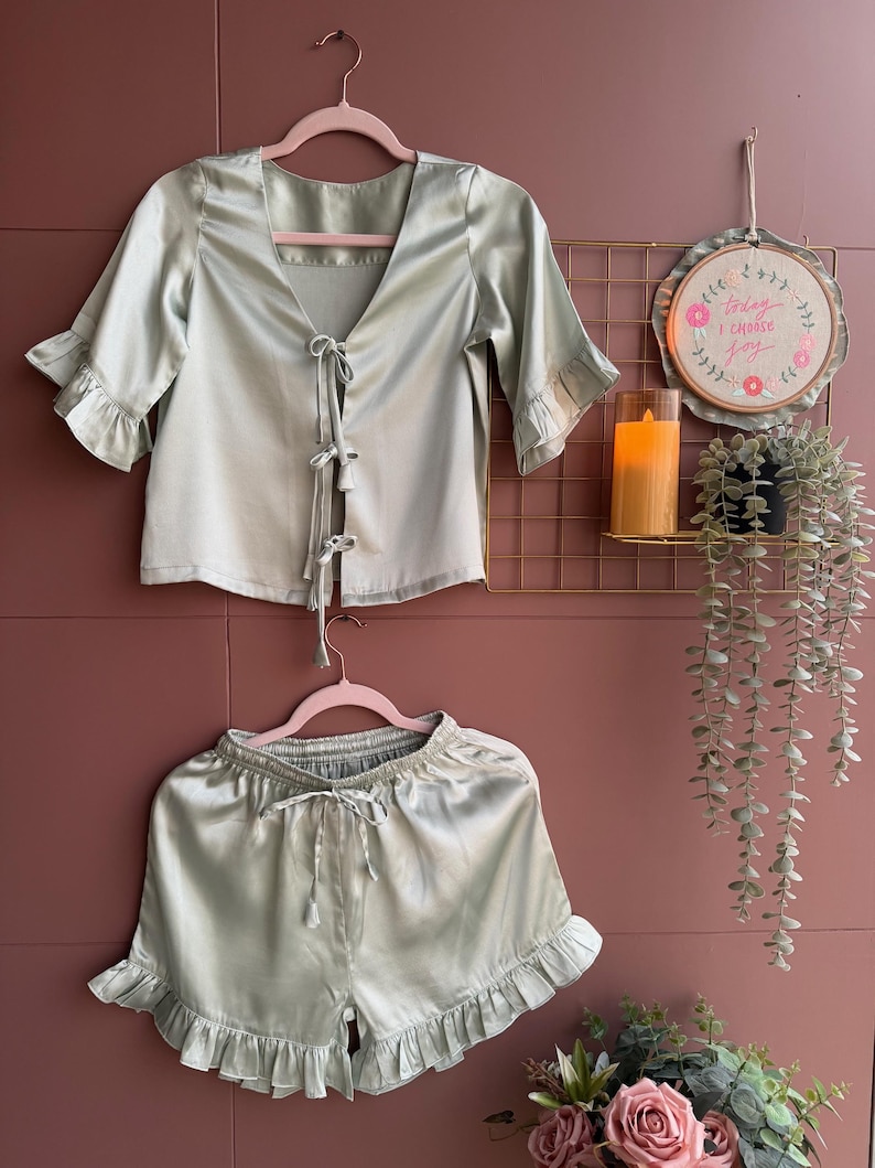 May include: A pale green satin pyjama set, comprising a top with a tie closure and frilled sleeves, and matching shorts with a drawstring waist and frilled hem. The set is displayed on pink hangers against a pink wall.