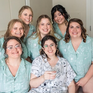 Puede incluir: Siete mujeres con pijamas a juego de color azul claro con estampado de gato blanco. Sonríen y miran a la cámara. Una mujer sostiene una copa de vino.