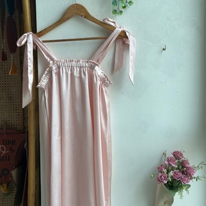May include: A light pink satin dress with thin straps that tie into bows at the shoulders. The dress is hanging on a wooden hanger against a white wall.