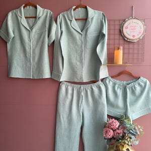 May include: A set of mint green linen pajamas, including a short-sleeved shirt, a long-sleeved shirt, pants, and shorts. Each piece has white piping and is displayed on a wooden hanger. The pajamas are arranged against a pink background.