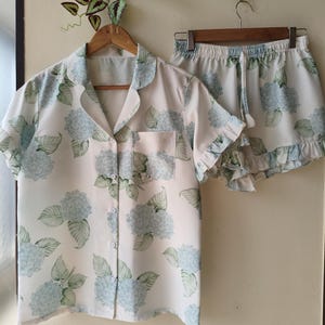 May include: A matching pajama set featuring a short-sleeved button-down shirt and shorts. The set is white with a blue and green floral pattern. The shorts have a ruffled hem and drawstring waist. The shirt has a chest pocket.