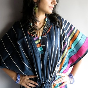 May include: A navy blue kaftan dress with vertical white stripes and colorful striped sleeves. The dress features a V-neck and a drawstring waist. A beaded necklace and blue bracelets complete the look.