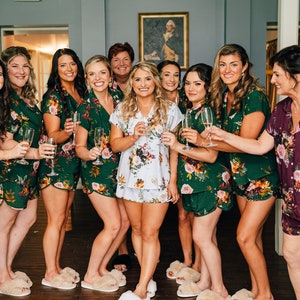Può includere: Un gruppo di donne in pigiami con stampa floreale abbinati, alcune con calici di champagne. I pigiami sono verdi, bianchi e viola con un motivo floreale. Le donne sorridono e sembrano festeggiare.