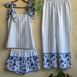 Op de afbeelding: Een driedelige pyjamaset in wit met blauwe verticale strepen. De set bevat een tanktop met strikbandjes, een korte broek en een broek. De korte broek en de broek hebben een blauwe bloemenrand. De tanktop heeft blauwe bloemenaccenten op de bandjes.
