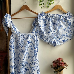 Op de afbeelding: Een witte pyjamaset met een blauw bloemenpatroon met vogels. Het topje heeft dunne bandjes en een vierkante halslijn. De shorts hebben een elastische tailleband.