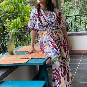 Puede incluir: Vestido caftán blanco con estampado ikat colorido en rojo, azul y amarillo. El vestido tiene cuello en V y cintura con cordón. Se usa con zapatos naranjas. El fondo incluye una mesa azul y vegetación verde. El vestido mide unos 150 cm de largo.