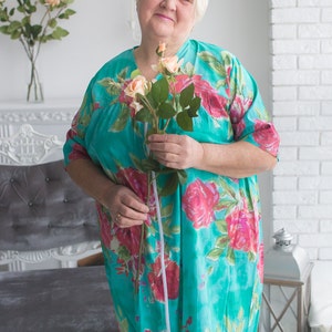 May include: A woman in a turquoise floral dress holds a bouquet of pink roses. She is standing in front of a white wall and a gray couch.