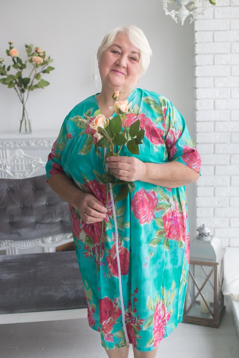 May include: A woman in a turquoise floral dress holds a bouquet of pink roses. She is standing in front of a white wall and a gray couch.