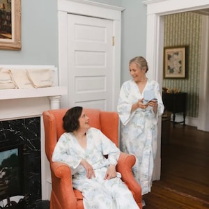 May include: Two people wearing white robes with blue floral patterns are standing in a room. The person on the left is sitting in a bright orange armchair. The person on the right is holding a small blue box.