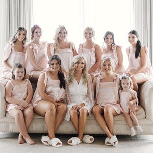 May include: A group of bridesmaids in pink satin pajamas pose for a photo on a white couch. The bride is in the center wearing a white robe with pearl embellishments.
