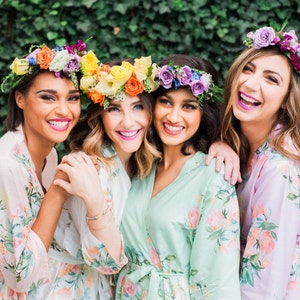May include: Four women wearing floral robes and flower crowns smile for a photo. The robes are in shades of pink, green, and purple with floral patterns. The flower crowns are made with a variety of colorful flowers.