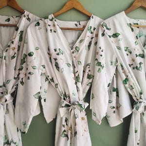 May include: Three white robes with a floral pattern of green leaves and white flowers. The robes have a V-neck and long sleeves. The robes are tied at the waist with a sash.