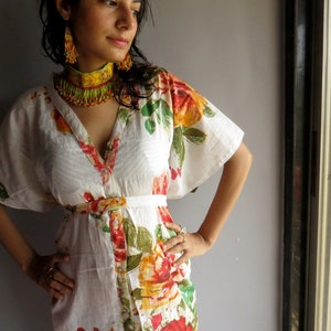 Op de afbeelding: Een witte maxi-jurk met een bloemenprint, met rode, gele en groene bloemen. De jurk heeft een knoopsluiting aan de voorkant en een riem. Het model draagt een gele en oranje kralenketting en bijpassende oorbellen.