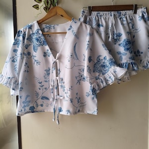 Op de afbeelding: Witte pyjamaset met een blauw bloemenpatroon. De set bevat een top met korte mouwen, een V-hals en een striksluiting, en een bijpassende short met een tailleband met trekkoord en een gefranjerde zoom. Het bloemmotief toont rozen.