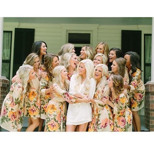 May include: A group of women in matching floral robes pose for a photograph. The women are smiling and laughing, and they are all wearing white robes with a floral pattern. The robes have long sleeves and a sash that ties at the waist. The women are standing in front of a white building with a porch.