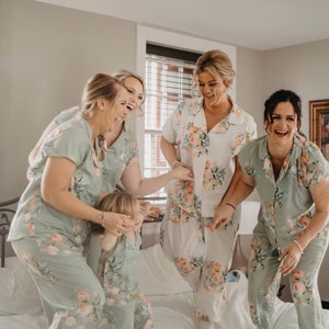 May include: Four women and a young girl wearing matching floral print pajamas are laughing and jumping on a bed. The pajamas are a light green color with a floral pattern in shades of pink and white.