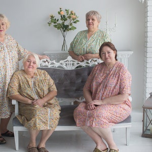 May include: Four women wearing floral print dresses pose for a photo. Two women are sitting on a white couch with gray upholstery. The other two women are standing behind the couch. The women are all smiling.