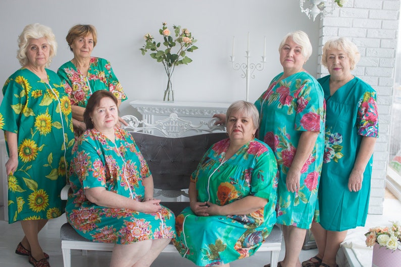 May include: Five women wearing teal floral print nightgowns. The women are standing and sitting in front of a white wall with a white fireplace mantel and a vase of flowers.