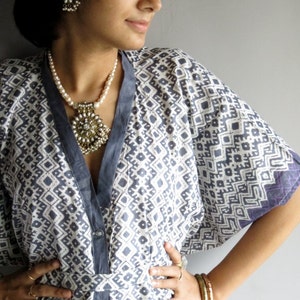 Puede incluir: Bata tipo kimono estampada en blanco y gris, con ribete y puños en gris oscuro. La bata presenta un diseño geométrico y se combina con un collar y pendientes de perlas. Anillos y pulseras doradas adornan el brazo.