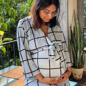 Puede incluir: Una mujer con una bata de color azul claro y cuadros negros. La bata tiene un lazo en la cintura y es holgada. La mujer está embarazada y se está sujetando el vientre.