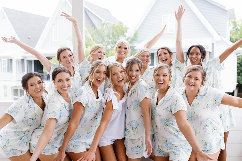 K&ouml;nnte beinhalten: Eine Gruppe von Frauen in passenden wei&szlig;en und blauen Blumenpyjamas posiert f&uuml;r ein Foto. Sie l&auml;cheln und lachen alle, einige von ihnen halten ihre Arme in die Luft. Die Frauen stehen vor einem wei&szlig;en Haus mit einer Veranda.