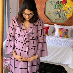 Puede incluir: Una persona embarazada lleva una bata de cuadros morados y negros con un cinturón de lazo. La bata tiene mangas largas y un escote en V. La persona está de pie frente a una cama con sábanas blancas y almohadas de colores.