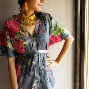 May include: A woman wearing a blue and floral print dress with a yellow and red beaded necklace and matching earrings. The dress has a button-up front and a belt.