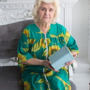 May include: A woman wearing a green and yellow floral dress is sitting on a gray chair. She is holding a blue and white patterned box.