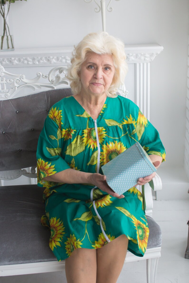 May include: A woman wearing a green and yellow floral dress is sitting on a gray chair. She is holding a blue and white patterned box.