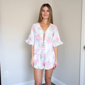 May include: White pajama set with a pink floral pattern. The top has a V-neck and ruffled sleeves, and the shorts have a ruffled hem. The set is tied in the front with a drawstring.
