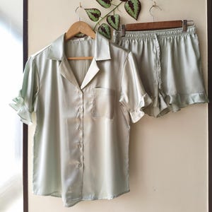 May include: A light green satin pajama set, including a button-down shirt with a chest pocket and ruffled sleeves, and matching shorts. The set is displayed on wooden hangers against a neutral wall, with decorative greenery.