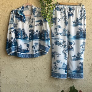 Op de afbeelding: Een witte en blauwe pyjamaset met een blauw en wit patroon. Het bovenstuk heeft een kraag en lange mouwen. De broek heeft een trekkoord in de taille en een wijde pijp.
