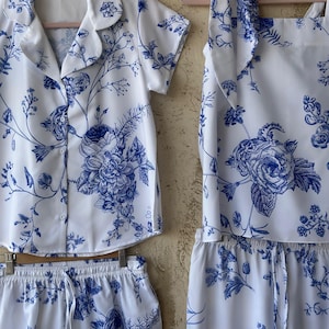 Op de afbeelding: Een witte pyjamaset met een blauwe bloemenprint. De set bevat een shirt met korte mouwen en een knoopsluiting, een broek met een trekkoord en een top zonder mouwen met een strik aan de hals.