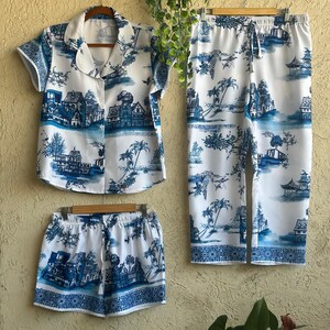 Op de afbeelding: Een witte pyjamaset met een blauw-wit patroon van huizen, palmbomen en bloemen. De set bevat een shirt met korte mouwen en een kraag en een broek.