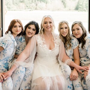 Op de afbeelding: Een bruid in een witte trouwjurk met bloemenkanten details, omringd door bruidsmeisjes in blauw-witte bloemenpyjama's. De vrouwen lachen en poseren voor een foto, wat een vrolijke sfeer creëert.